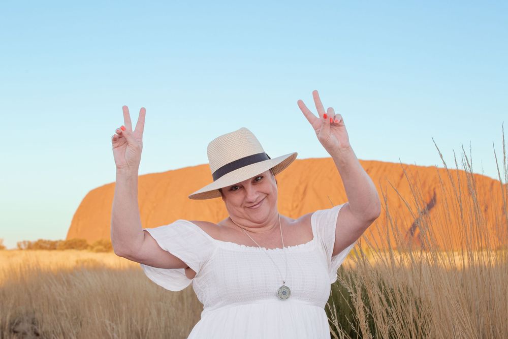 Someone wearing a white shirt and straw hat makes peace signs while posing in front of golden grass and blue sky.