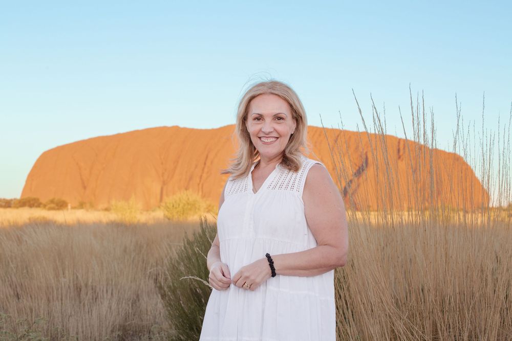 A woman in a white dress smiles while walking in a field of tall dried grass during sunset.