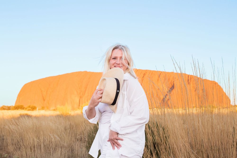 Someone in white clothing holds a hat while standing in front of Uluru at sunset in Australia's Northern Territory.