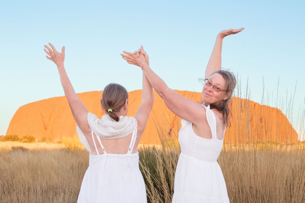 Two people in white dresses raise their arms joyfully in front of Uluru rock formation at sunset.