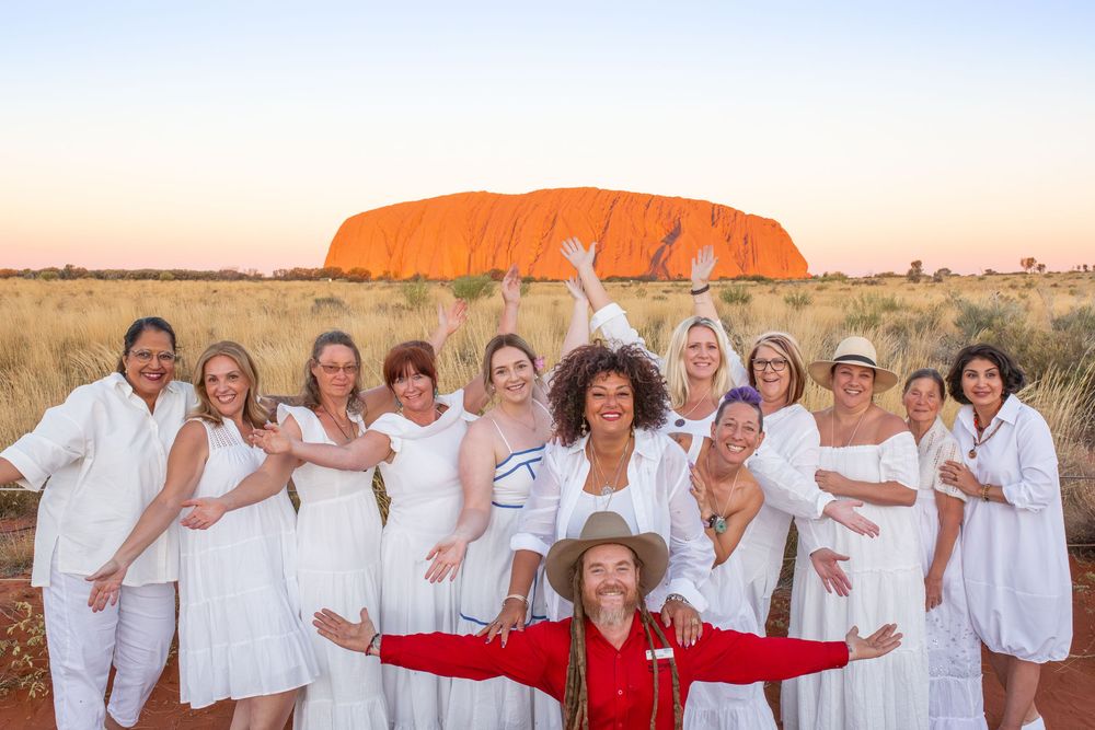 A group wearing white outfits pose in front of Uluru rock formation in Australia's outback during sunset.