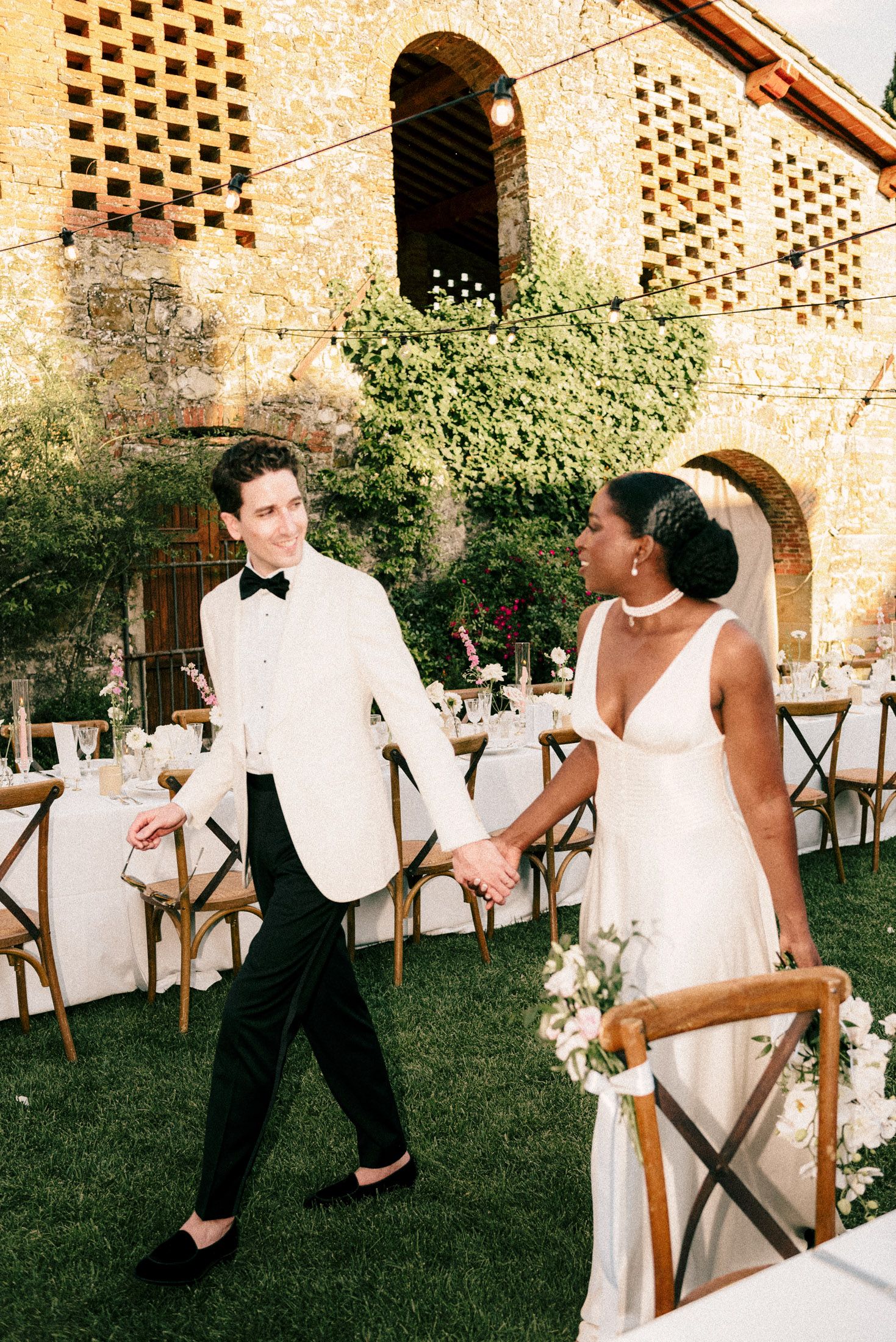 Elegant wedding couple holding hands at outdoor reception with stone building and rustic decor in background.