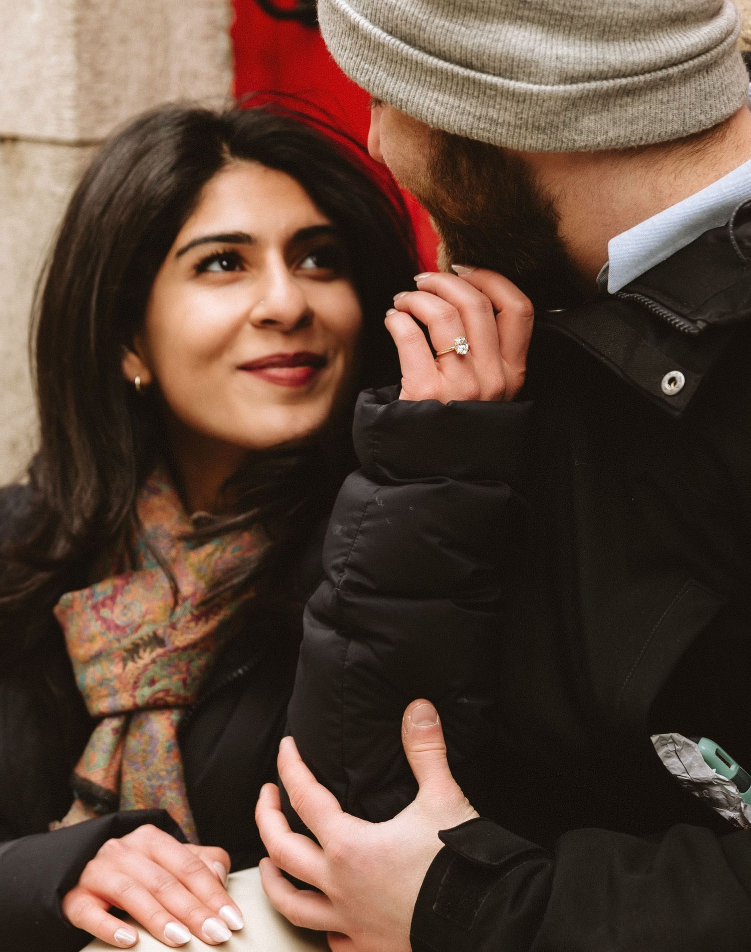 Intimate winter portrait of couple showing engagement ring against red wall background.