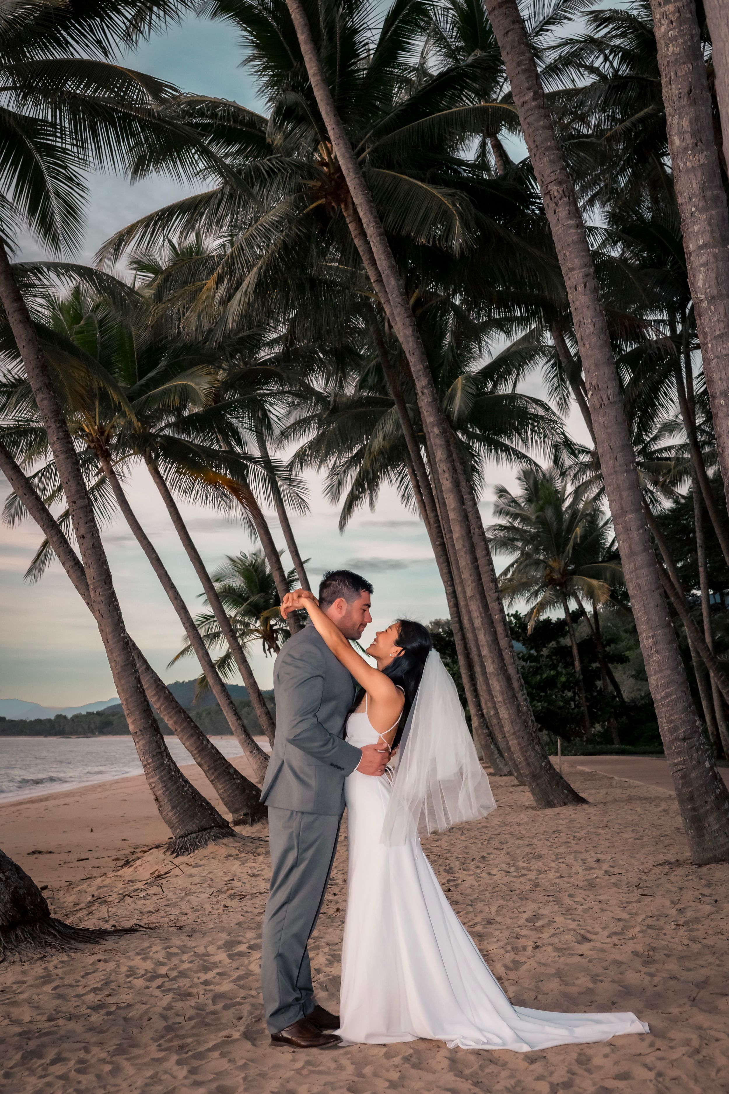 A couple embraces on a tropical beach at sunset surrounded by towering palm trees in Hawaii.