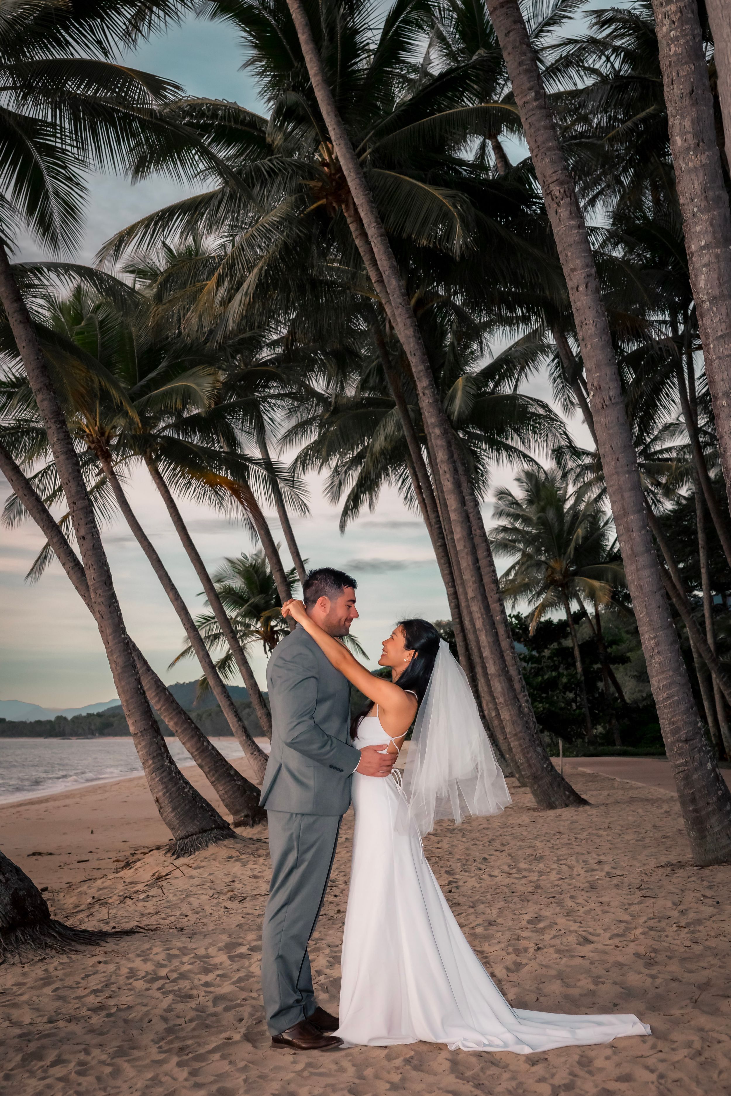 Newlywed couple shares romantic moment on a tropical beach at sunset surrounded by towering palm trees.