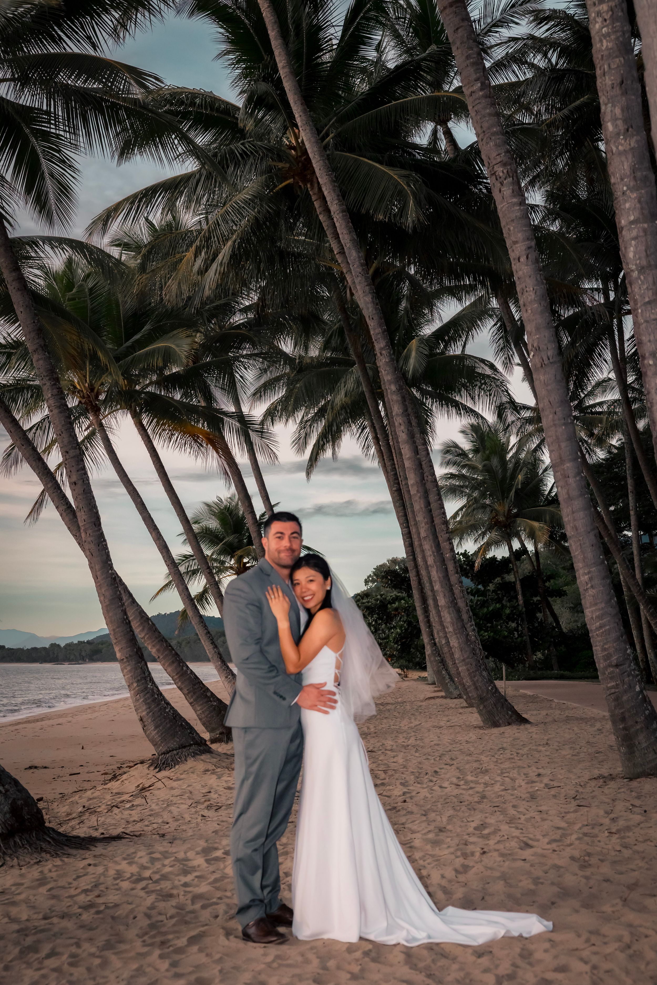 A romantic couple poses on a tropical beach at sunset surrounded by tall palm trees on the sandy shore.