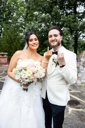 A newlywed couple in white attire pose outdoors with a bouquet of pink and white flowers on their wedding day.