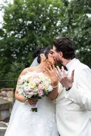 A romantic wedding scene outdoors shows a couple sharing a kiss while the bride holds a beautiful pink and white bouquet.