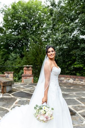 Person in a flowing white wedding dress poses with a bouquet against a stone wall and lush greenery backdrop.