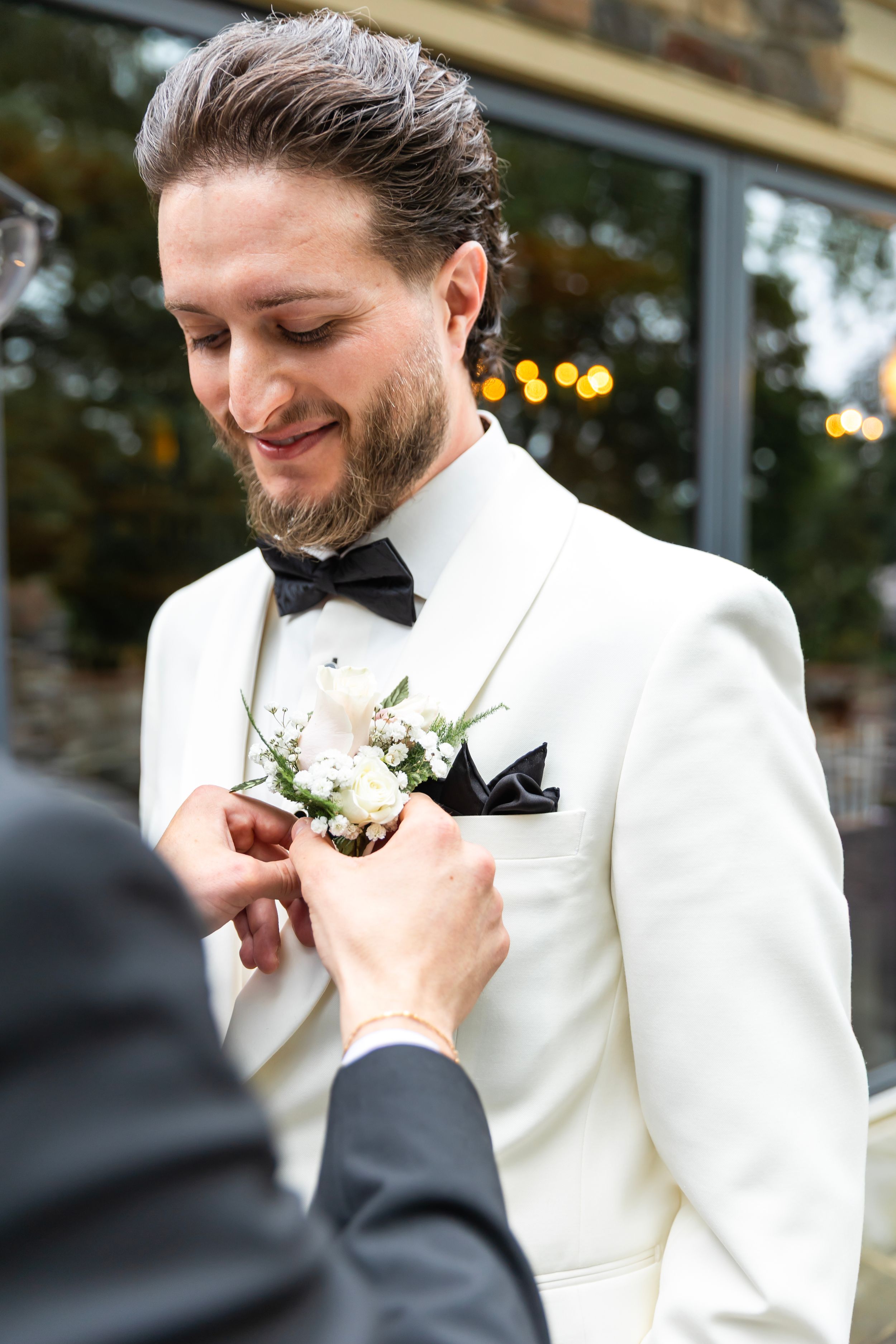 Someone helps pin a delicate white boutonniere to a groom's crisp white tuxedo jacket at an elegant wedding.NJ wedding photographer