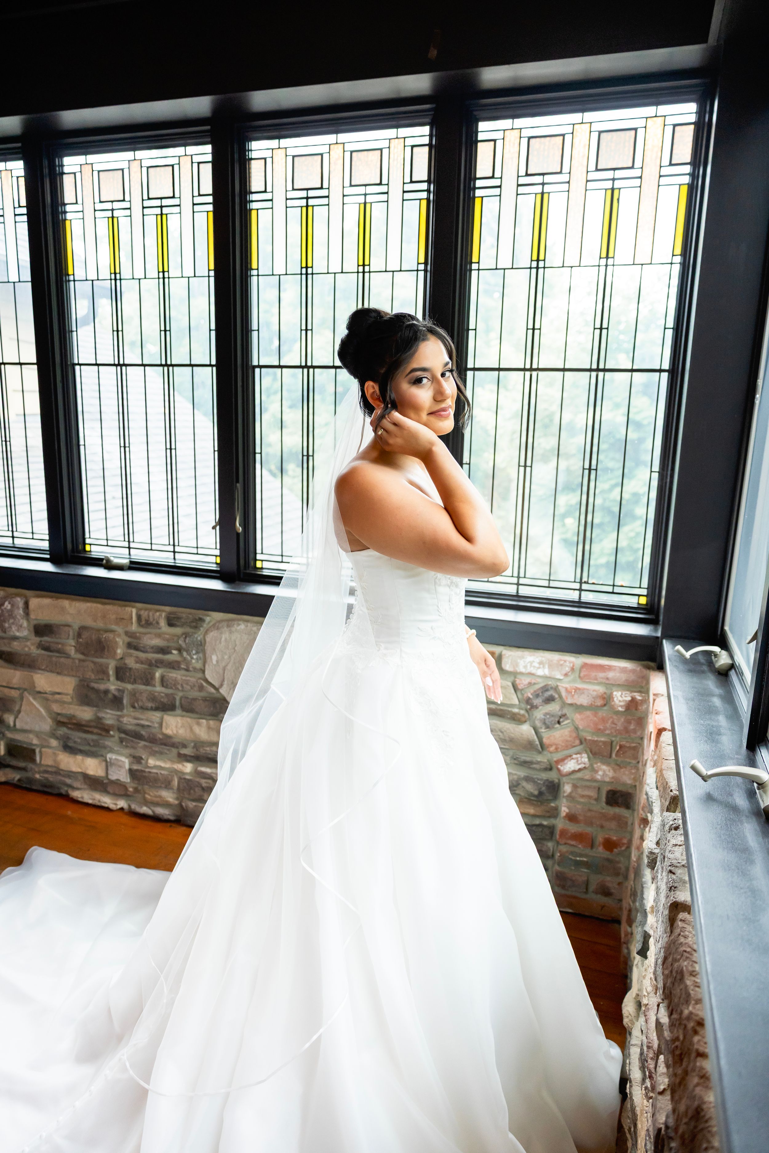 A bride in an elegant white strapless wedding gown poses by a window with decorative stained glass panels.