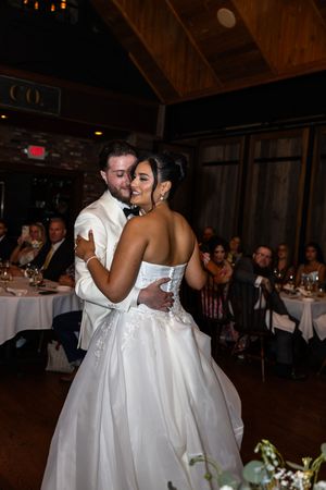 A couple shares their first dance as newlyweds at an elegant indoor reception with wooden beams and white table settings.