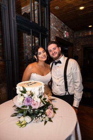 A wedding couple cuts their elegant white wedding cake adorned with purple and white flowers in a rustic venue.