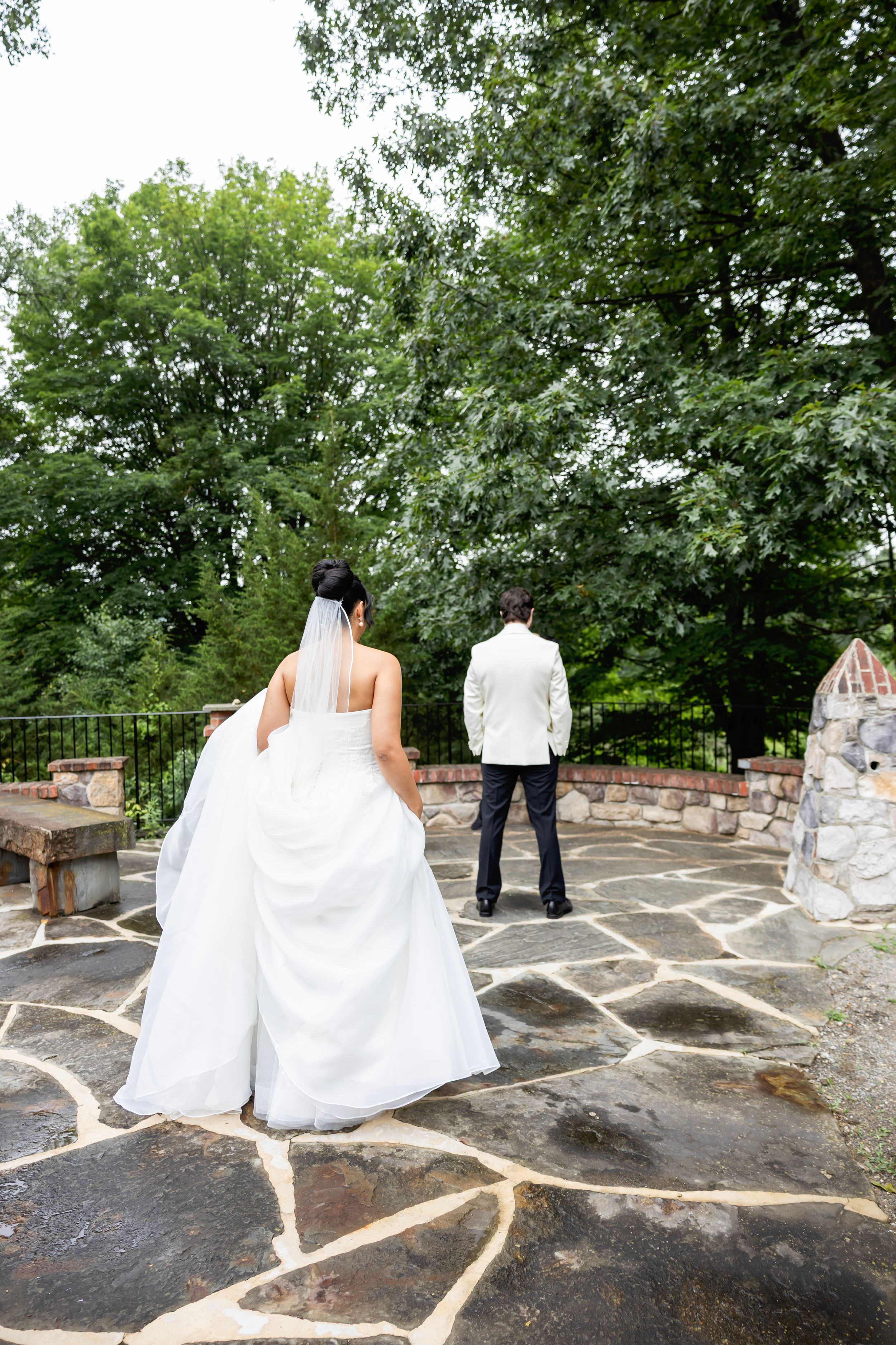 A bride and groom stand on a stone patio surrounded by lush green trees during an outdoor wedding ceremony.