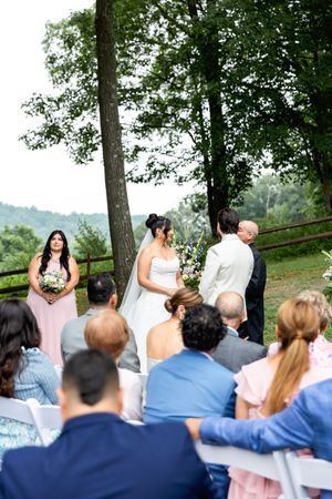 A wedding ceremony takes place outdoors with guests seated on a hillside overlooking scenic mountain views.