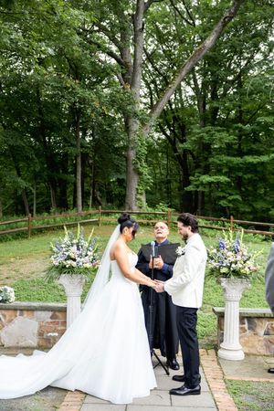 Wedding ceremony unfolds on an outdoor stone patio surrounded by lush green trees and elegant white floral arrangements.