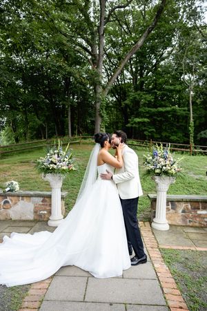 A romantic wedding ceremony with the couple sharing a kiss outdoors with decorative floral pillars and lush greenery.