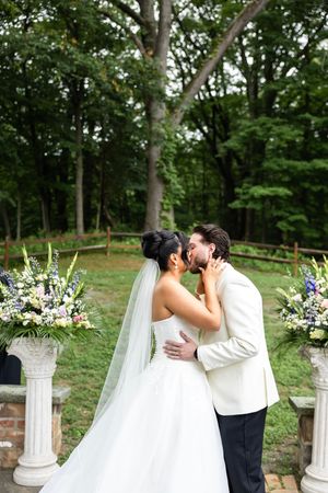 A romantic outdoor wedding moment with white columns and purple floral arrangements in a lush garden setting.