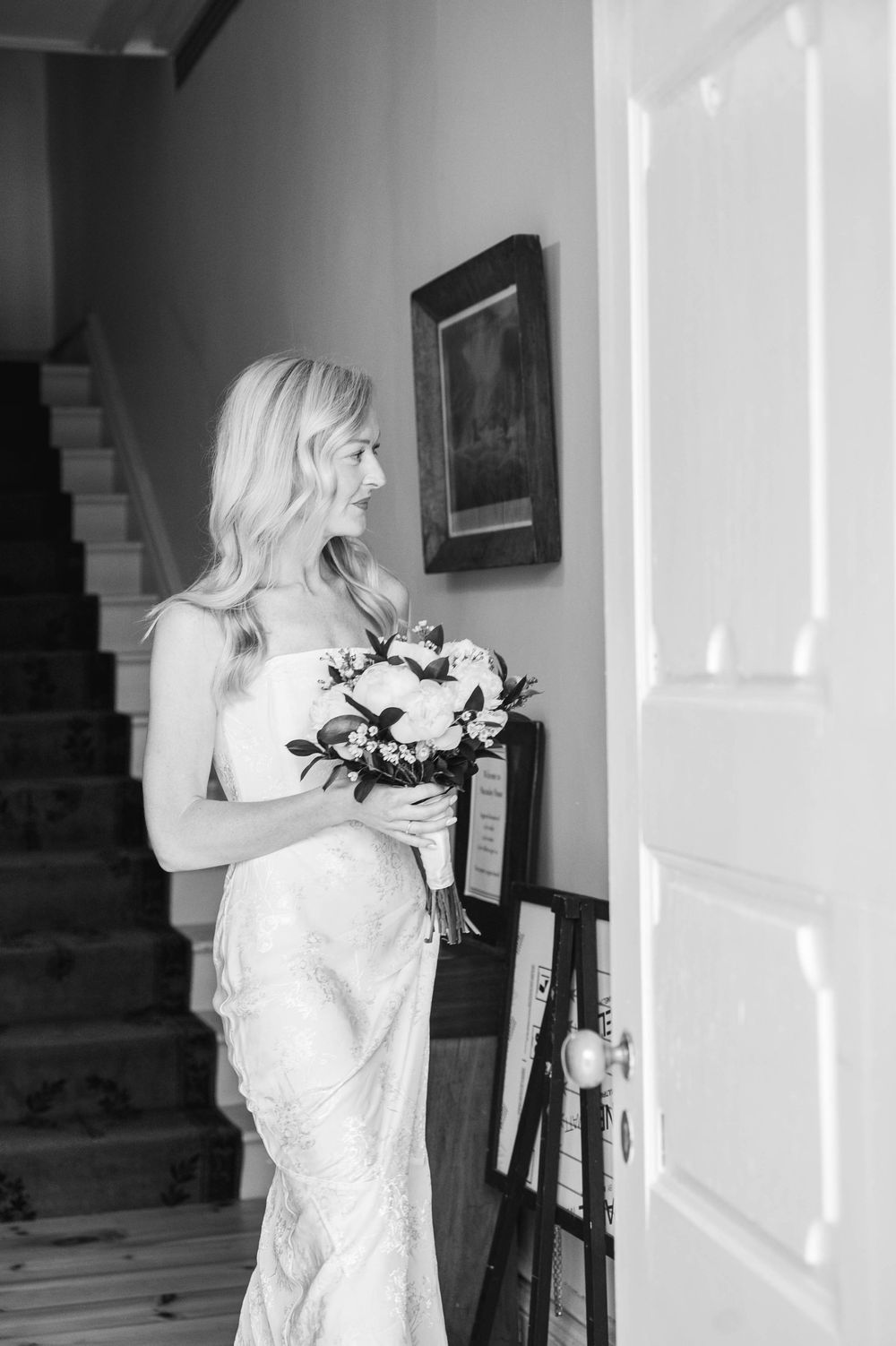 Bride in a fitted lace wedding gown holding a white bouquet beside a staircase in black and white.