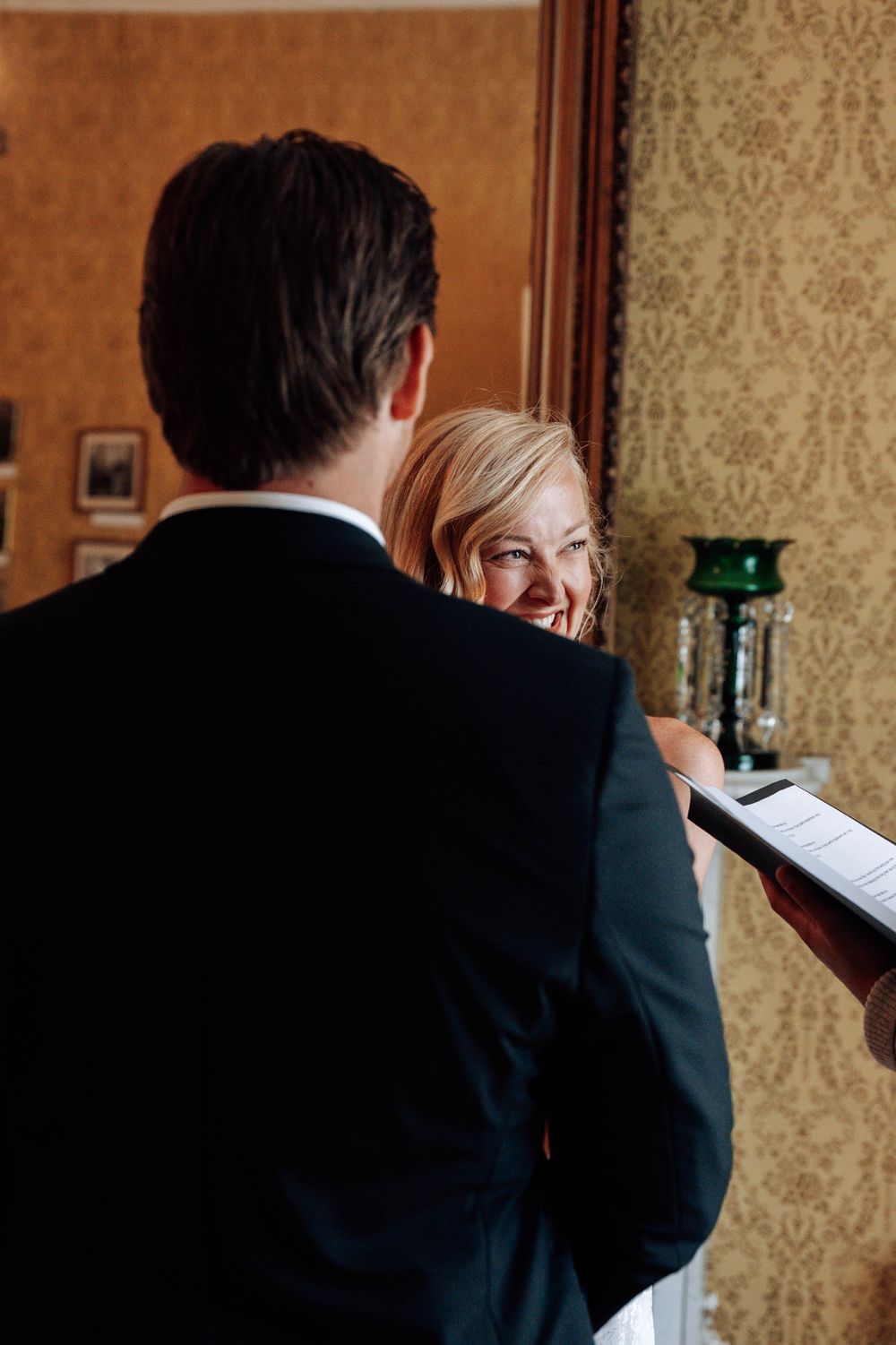 A couple shares a joyful moment during their wedding ceremony while reading vows in an elegant indoor setting.