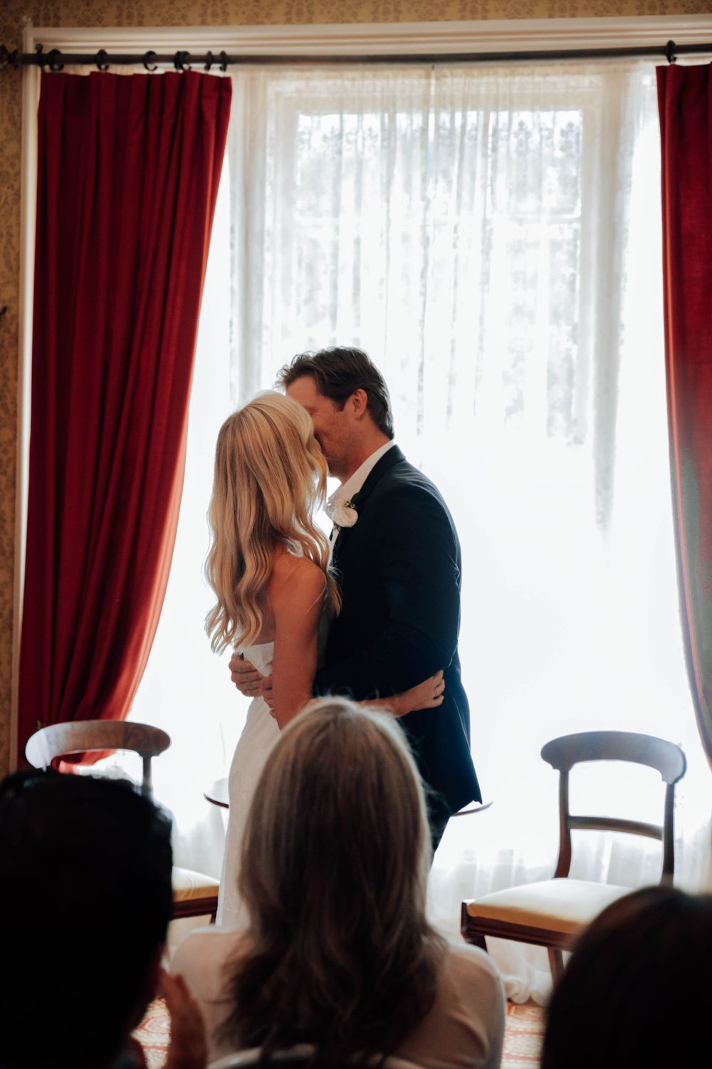 Silhouettes of wedding ceremony moments captured against bright window light with red curtains.