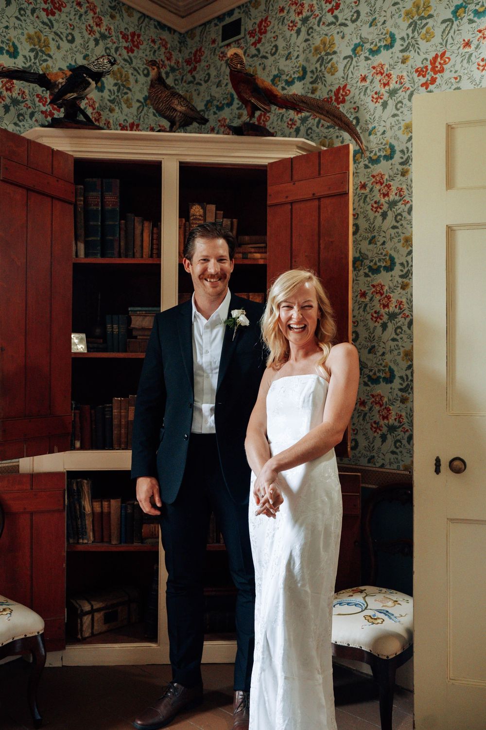 Newlyweds pose in a library room with floral wallpaper and wooden cabinets.