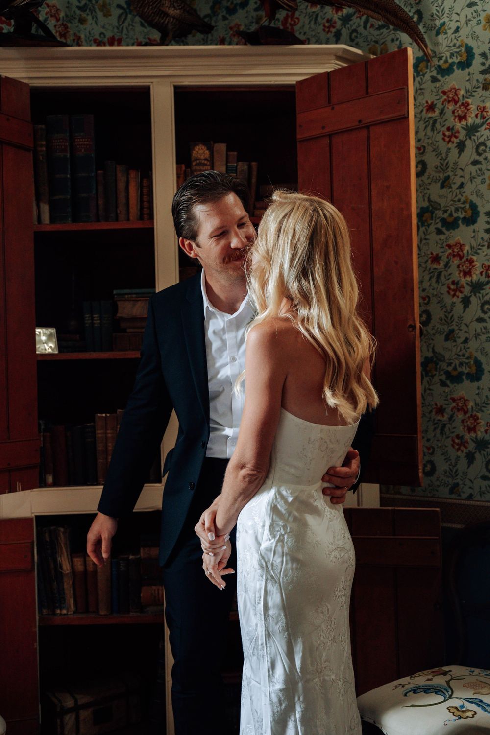 A couple shares intimate moments in an indoor setting with vintage wallpaper and wooden furniture.