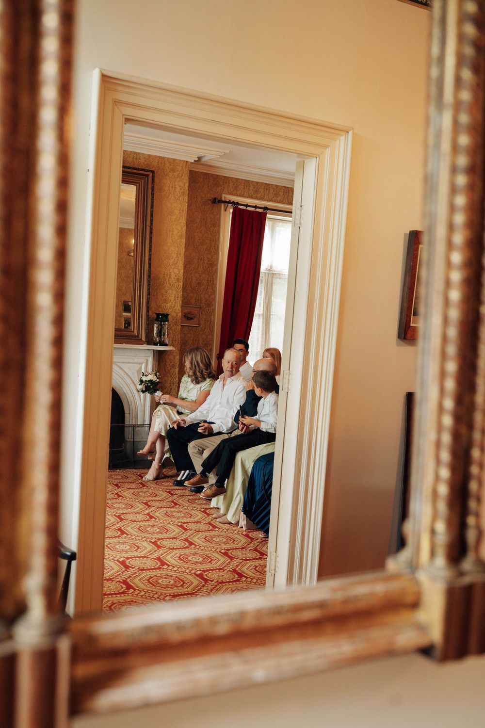 A mirror reflection shows wedding guests seated in an elegant room with red carpet.