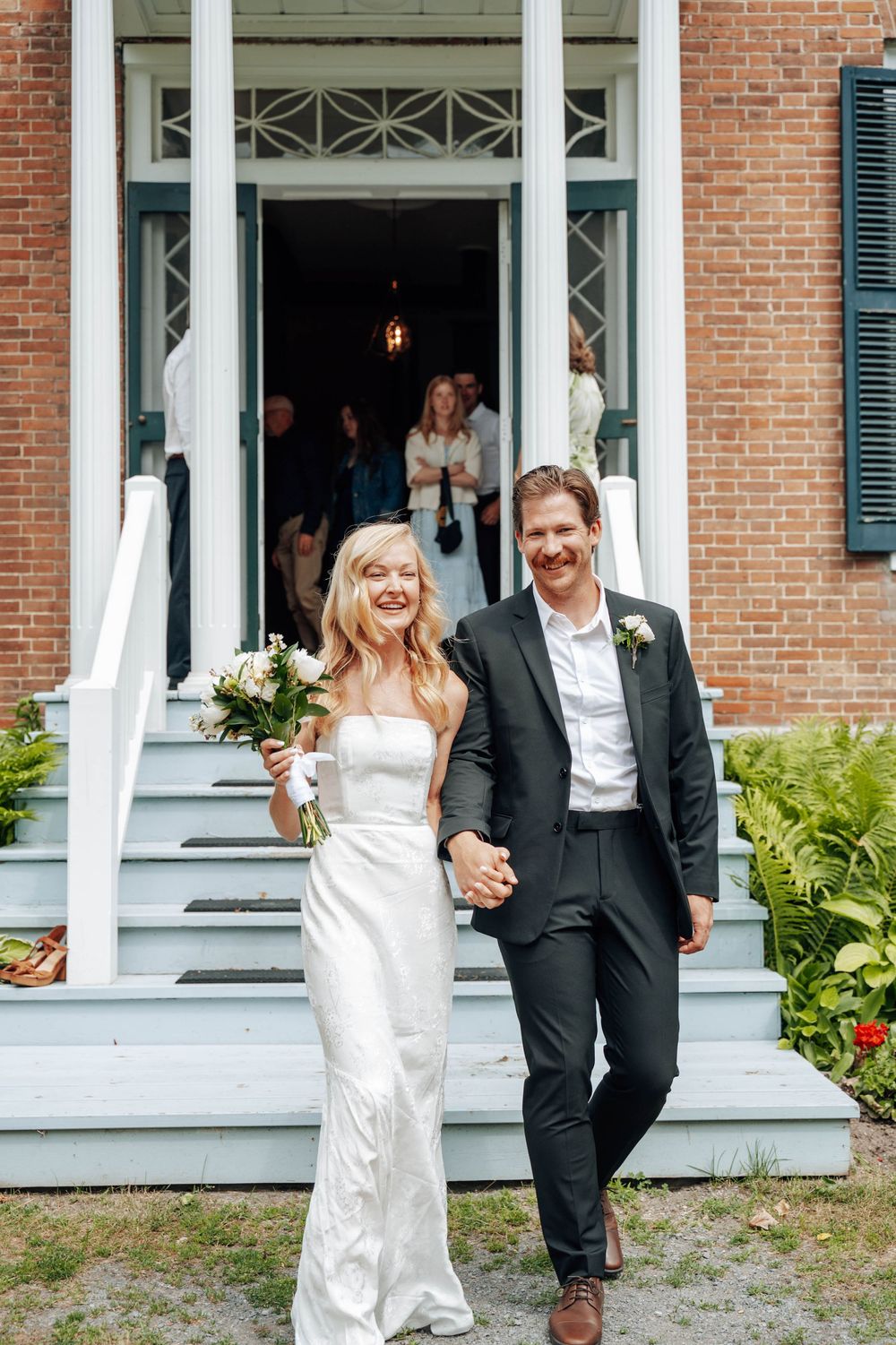 A newlywed couple exits a historic brick building with white columns and steps.