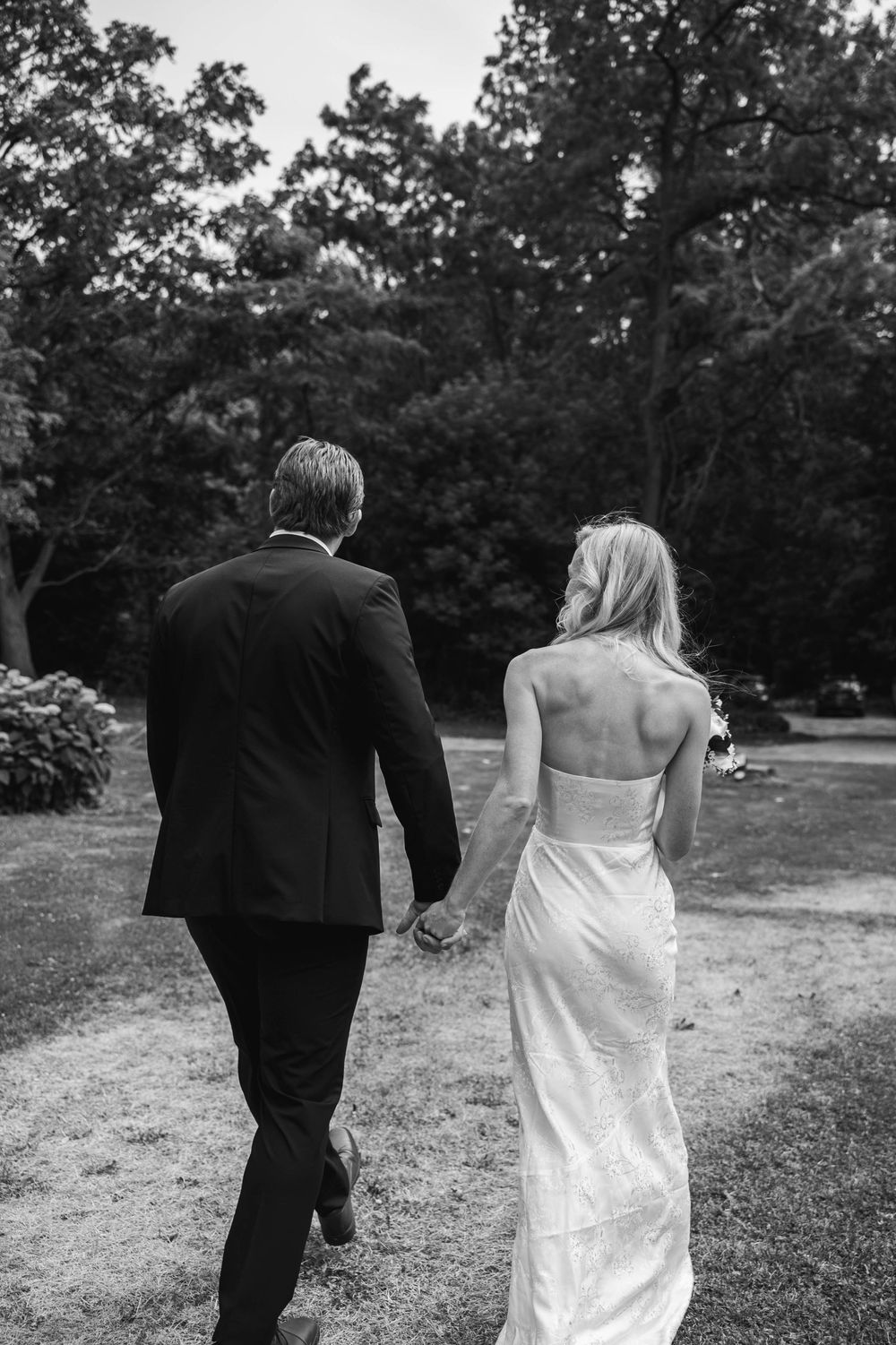 A couple in formal wedding attire walking away from the camera across a grassy field with trees in the background.