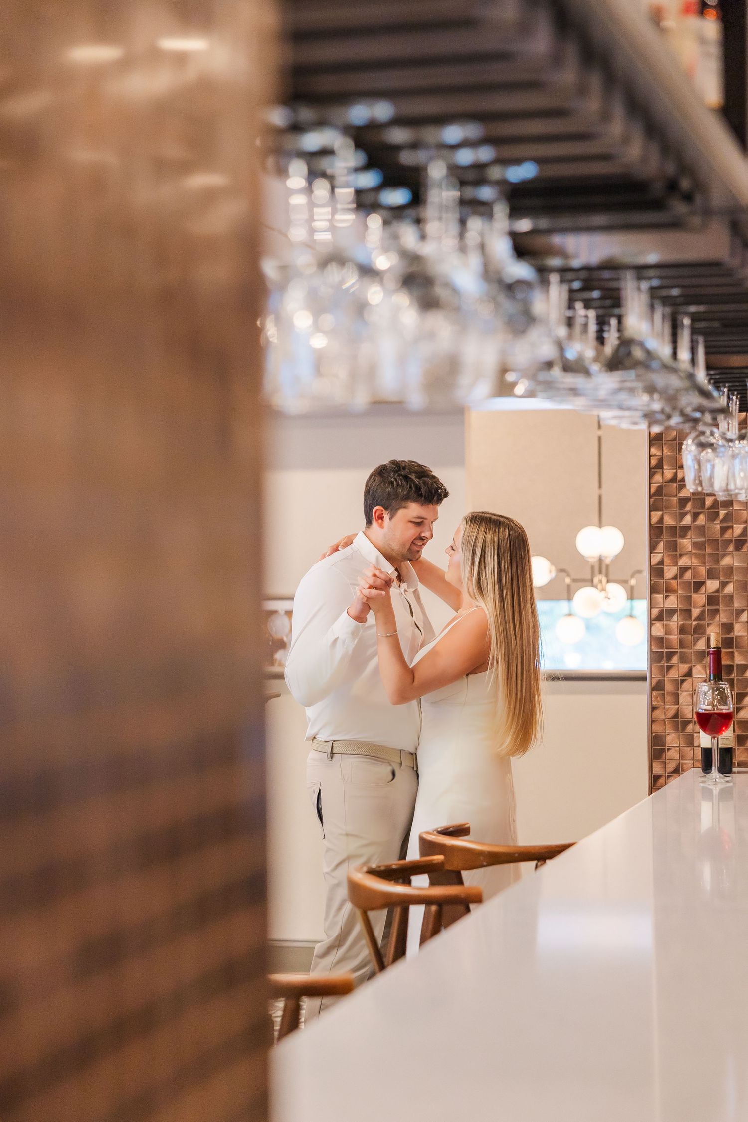 A romantic couple shares an intimate moment seated at a wine bar counter in a softly lit restaurant setting.