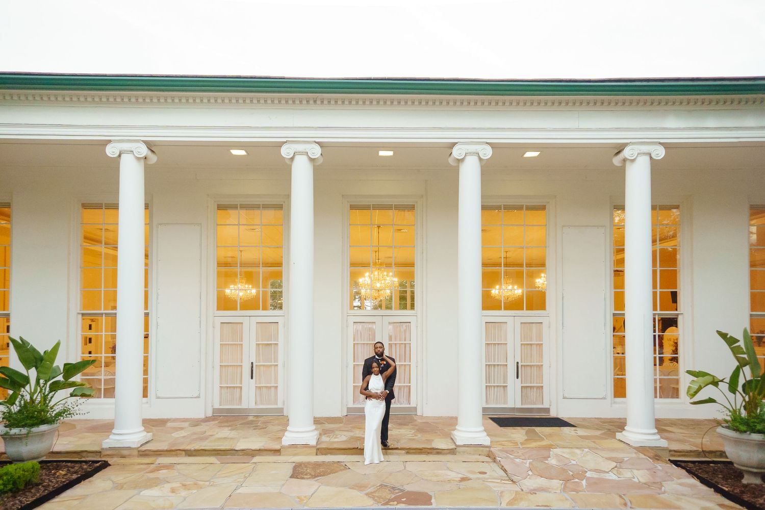 A couple stands embracing between white classical columns of an elegant venue with golden-lit windows.