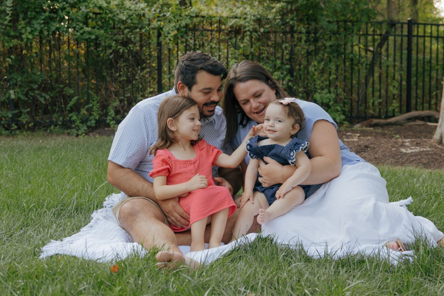 A family sits together on white blankets in a grassy outdoor setting during a casual photo session.