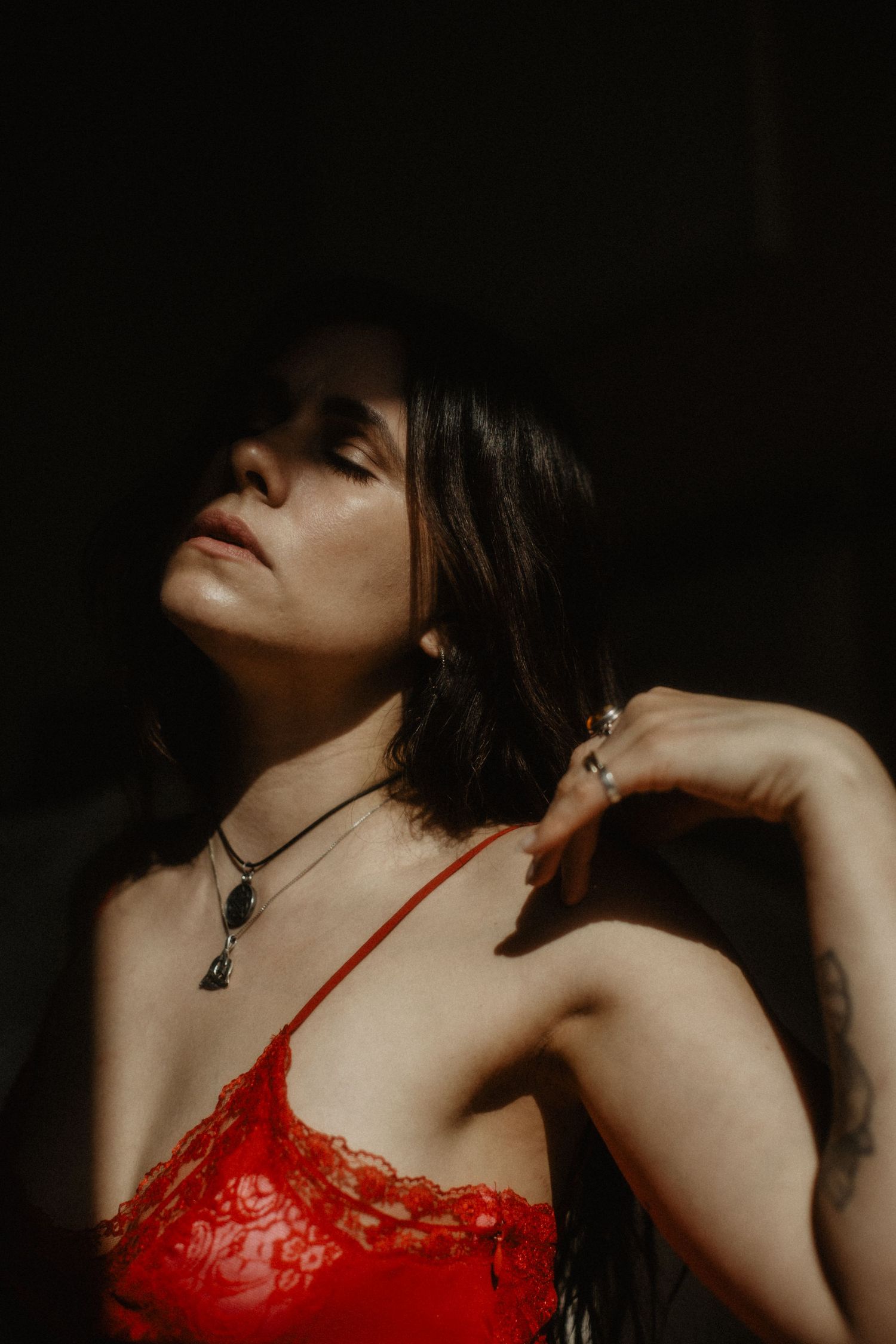 Moody portrait in dark lighting shows figure in red lace lingerie with necklace, head tilted back dramatically.