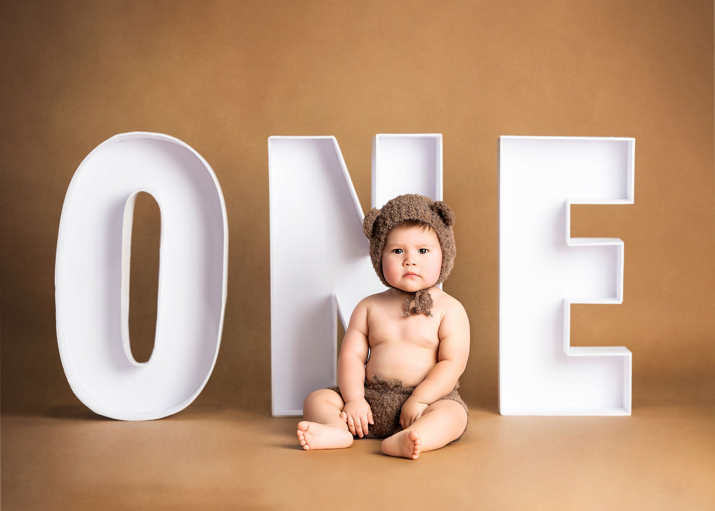 A small child sits between large white letters spelling out ONE against a brown backdrop.