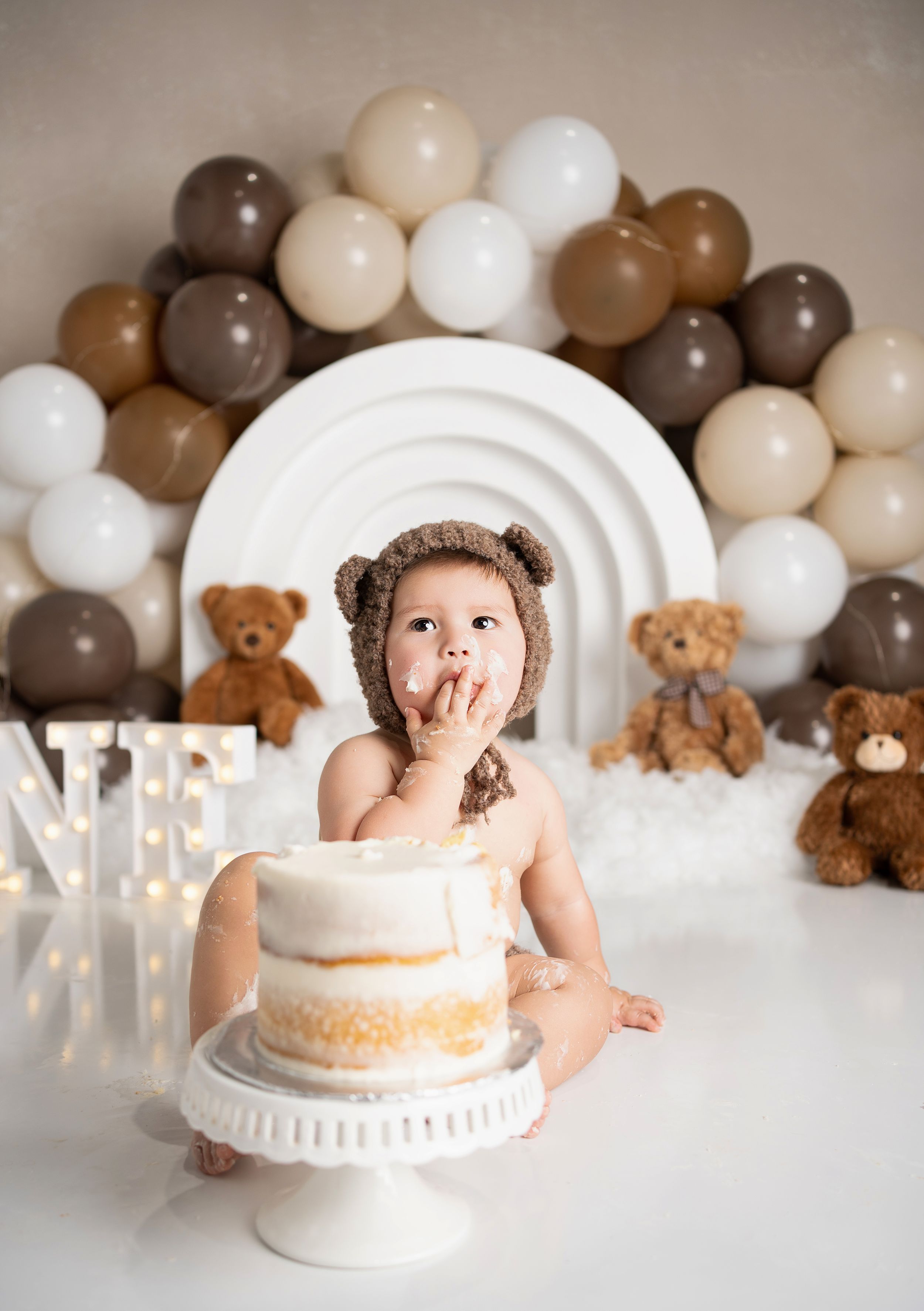 First birthday cake smash photo setup with brown and neutral balloon arch, teddy bears, and white cake on pedestal stand.