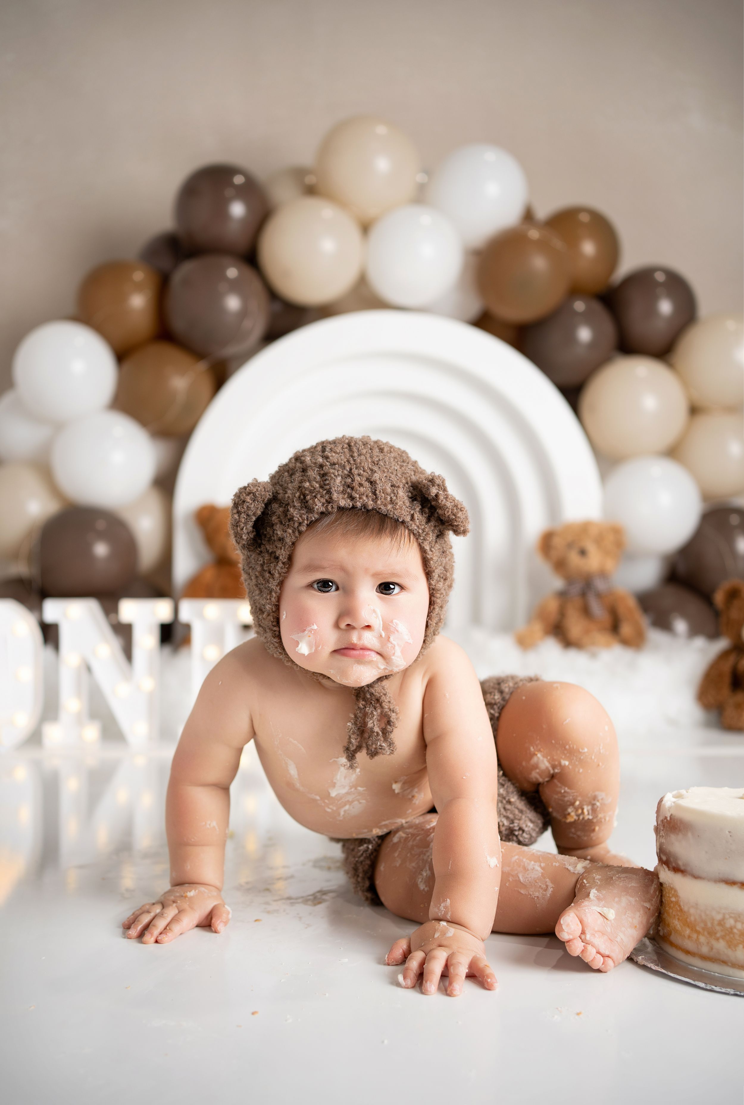 Festive first birthday photo setup with brown and beige balloon arch, teddy bear and cake smash props on white backdrop.