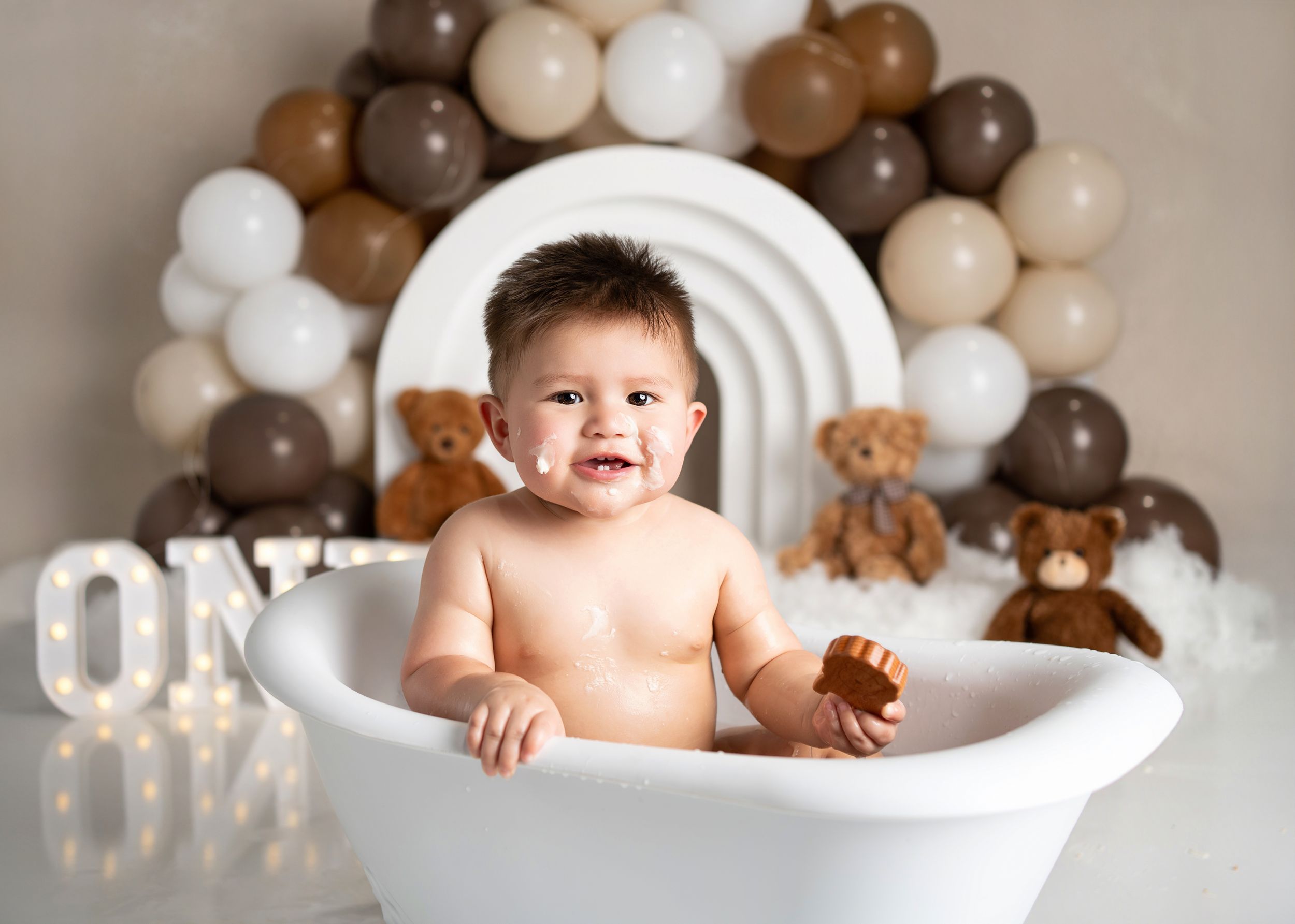 A baby sits in a white bathtub with brown and beige balloon arch and teddy bears for first birthday milestone photo.
