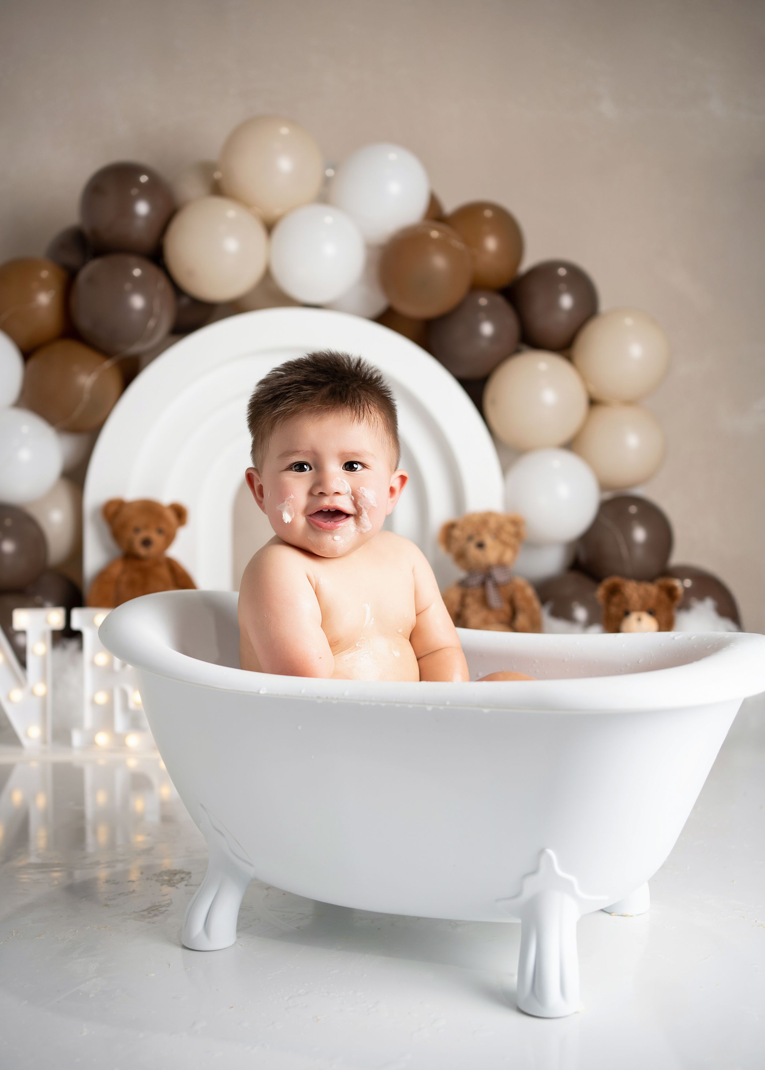 Baby sitting in  white bathtub with balloon arch and teddy bear decorations in neutral tones background.