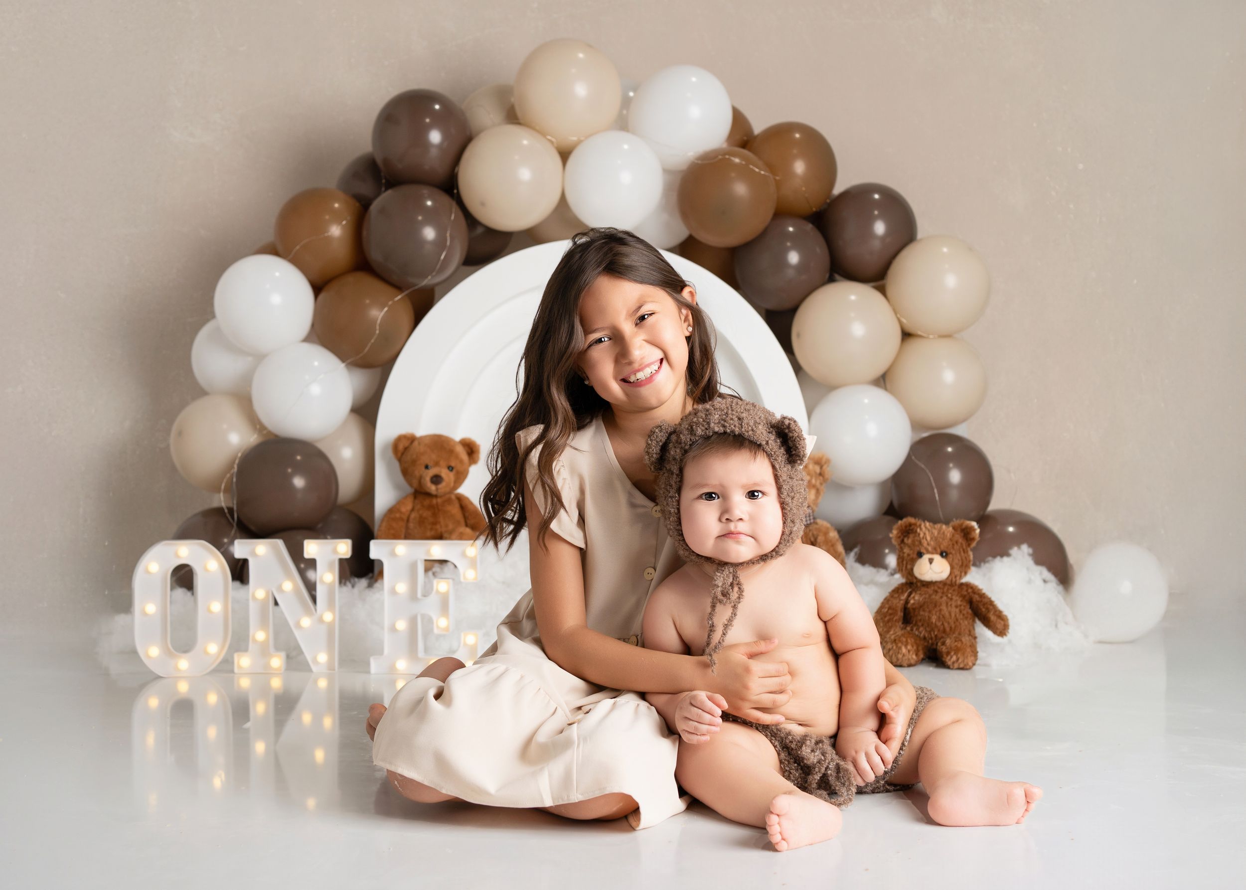 A baby and sister pose together during a studio photography session with neutral tones and balloons and teddy bears.