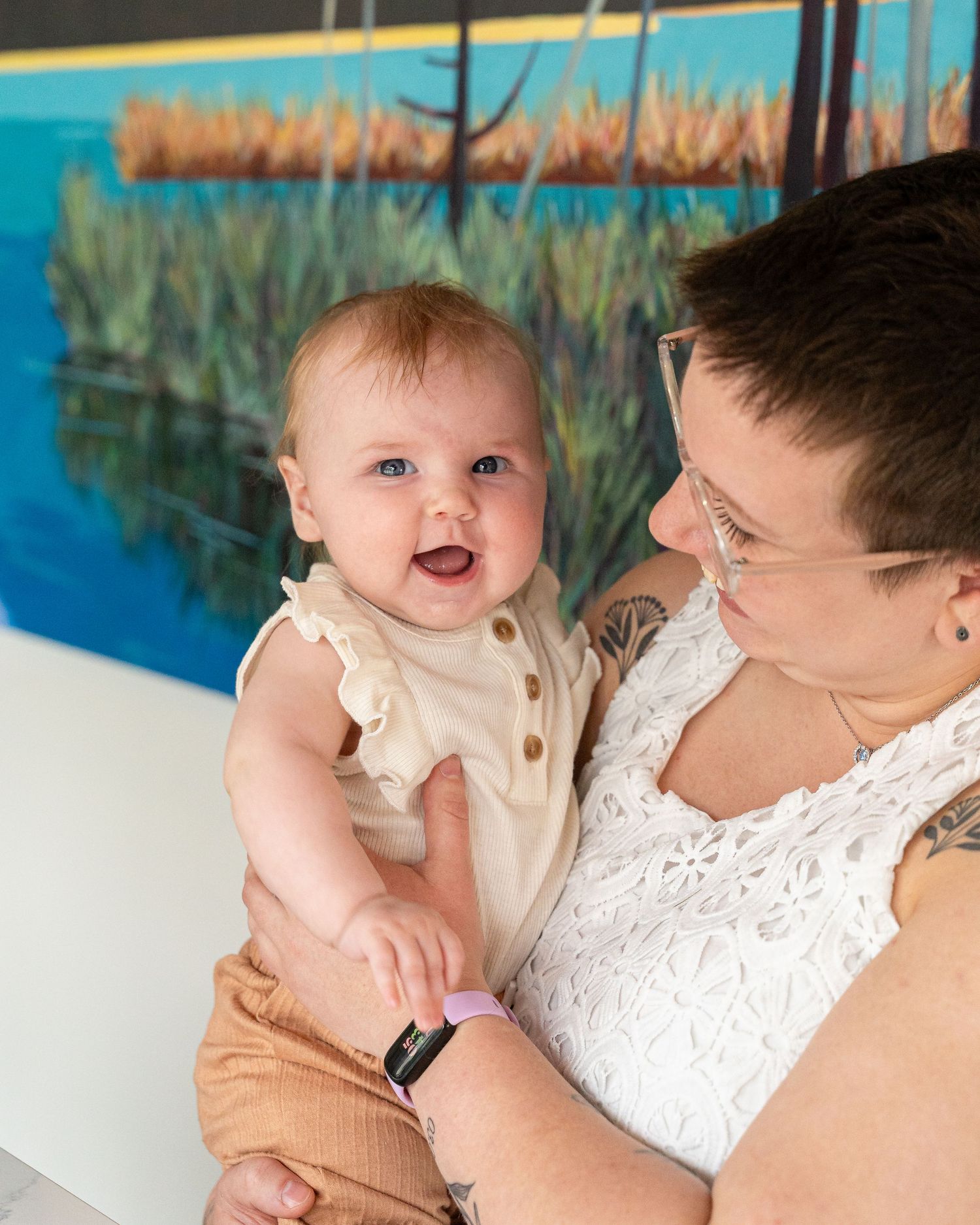 Two adults enjoying coffee at a table with a baby while sitting in front of a bookshelf and colorful wall mural.