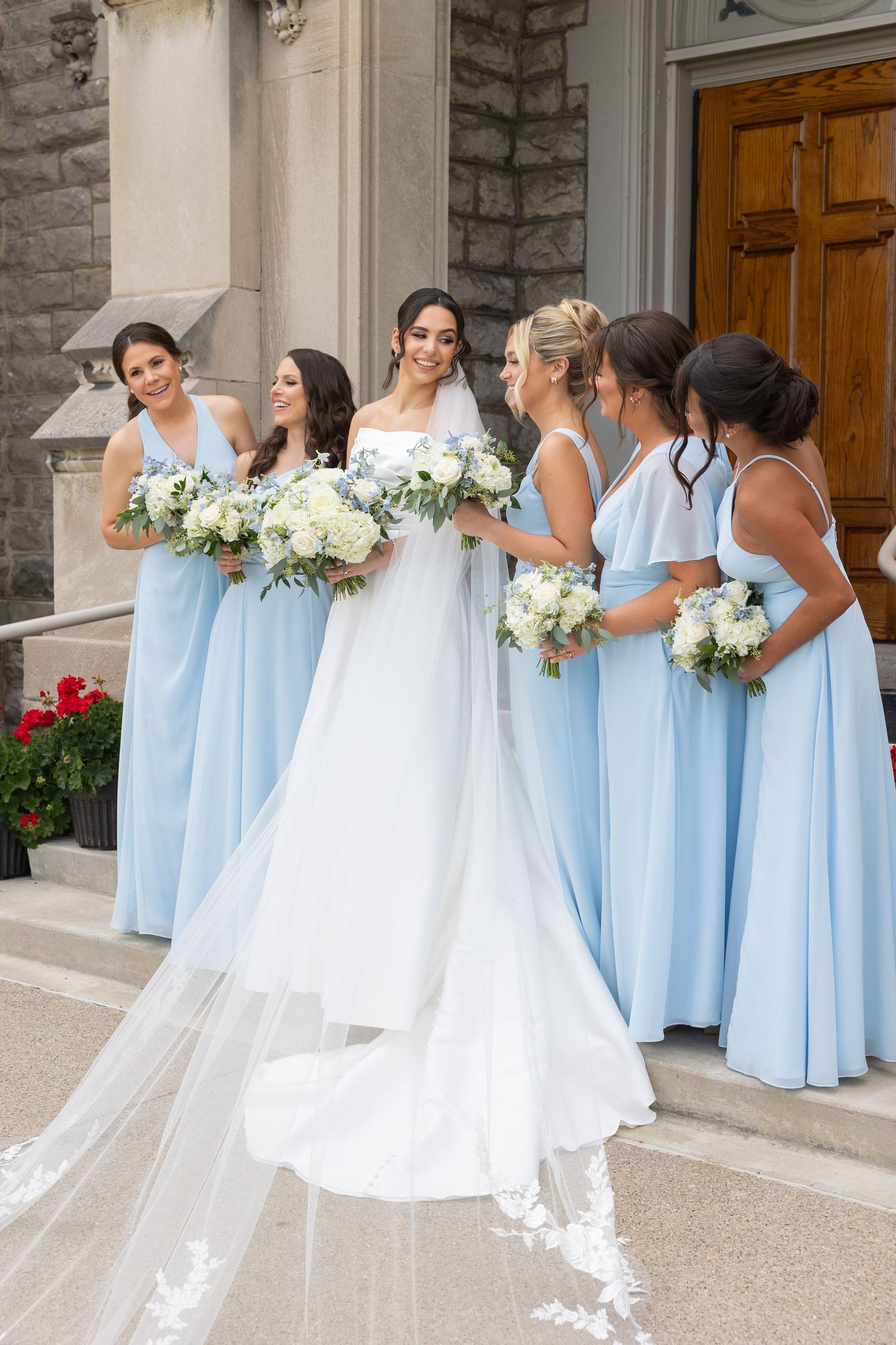A bridal party in light blue bridesmaid dresses holding white floral bouquets stands together on church steps. Rachel Martinez Photography