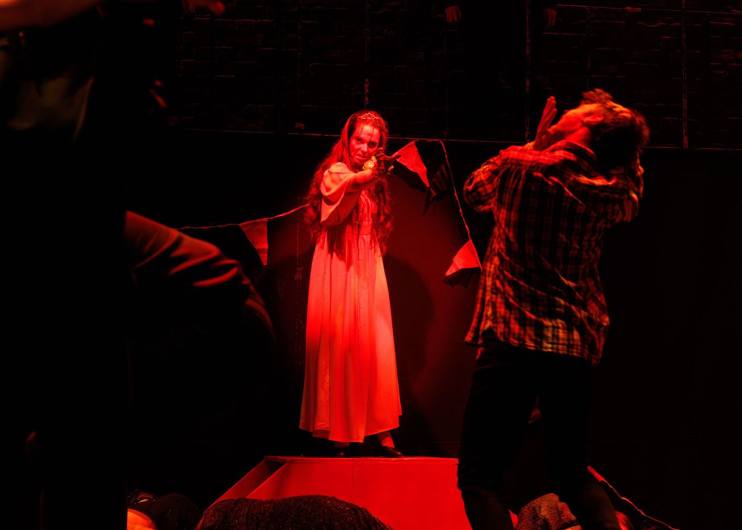 A dramatic scene bathed in red lighting shows performers in theatrical poses on a dark stage.