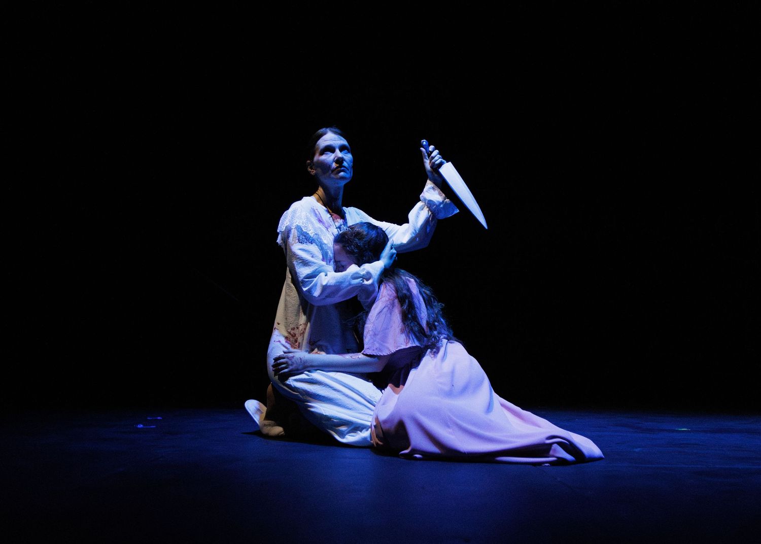 A stage performer lit in blue lighting kneels in white traditional costume while holding a curved blade during a dramatic scene.