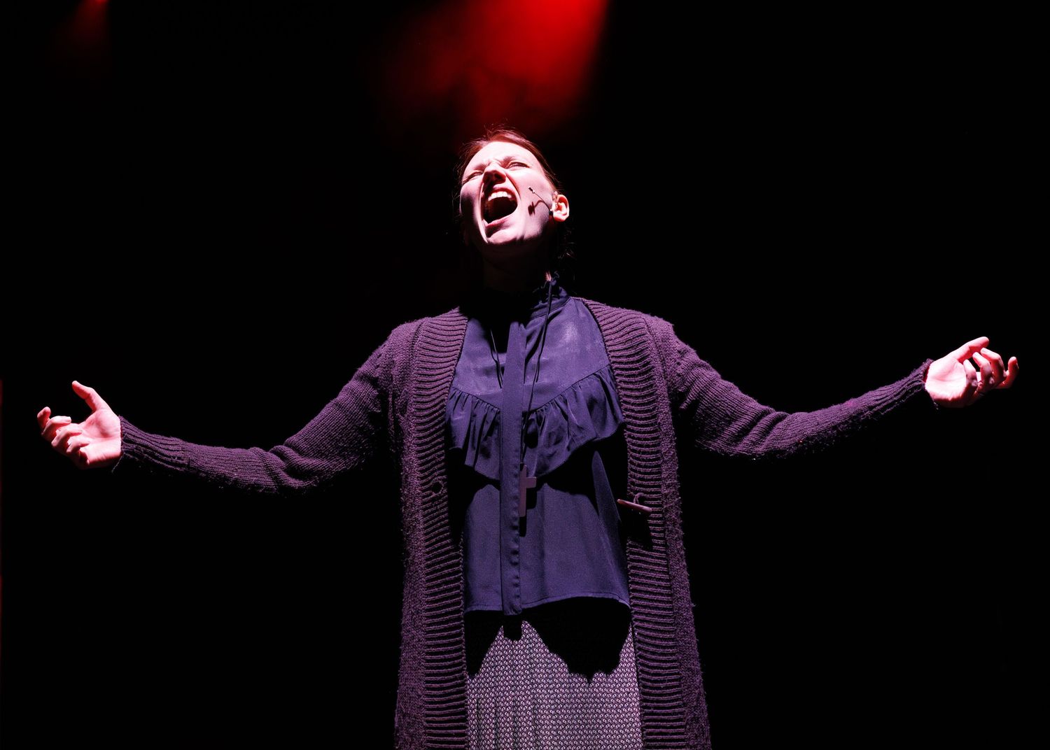 A performer in dark Victorian-style clothing stands with arms outstretched on a dimly lit stage with red lighting.