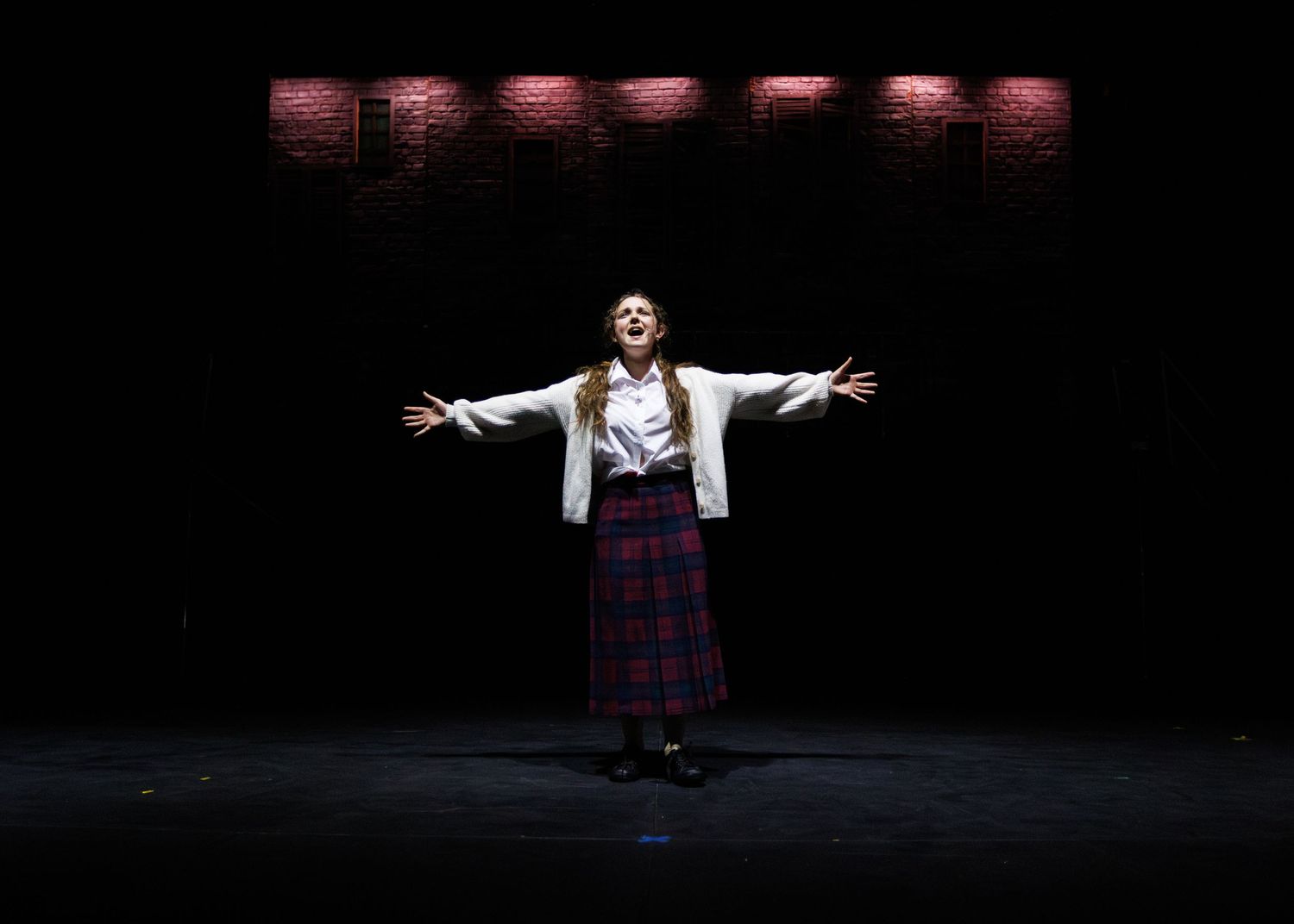 A performer stands with arms outstretched on a dark stage wearing a white jacket and plaid skirt under dramatic red lighting.