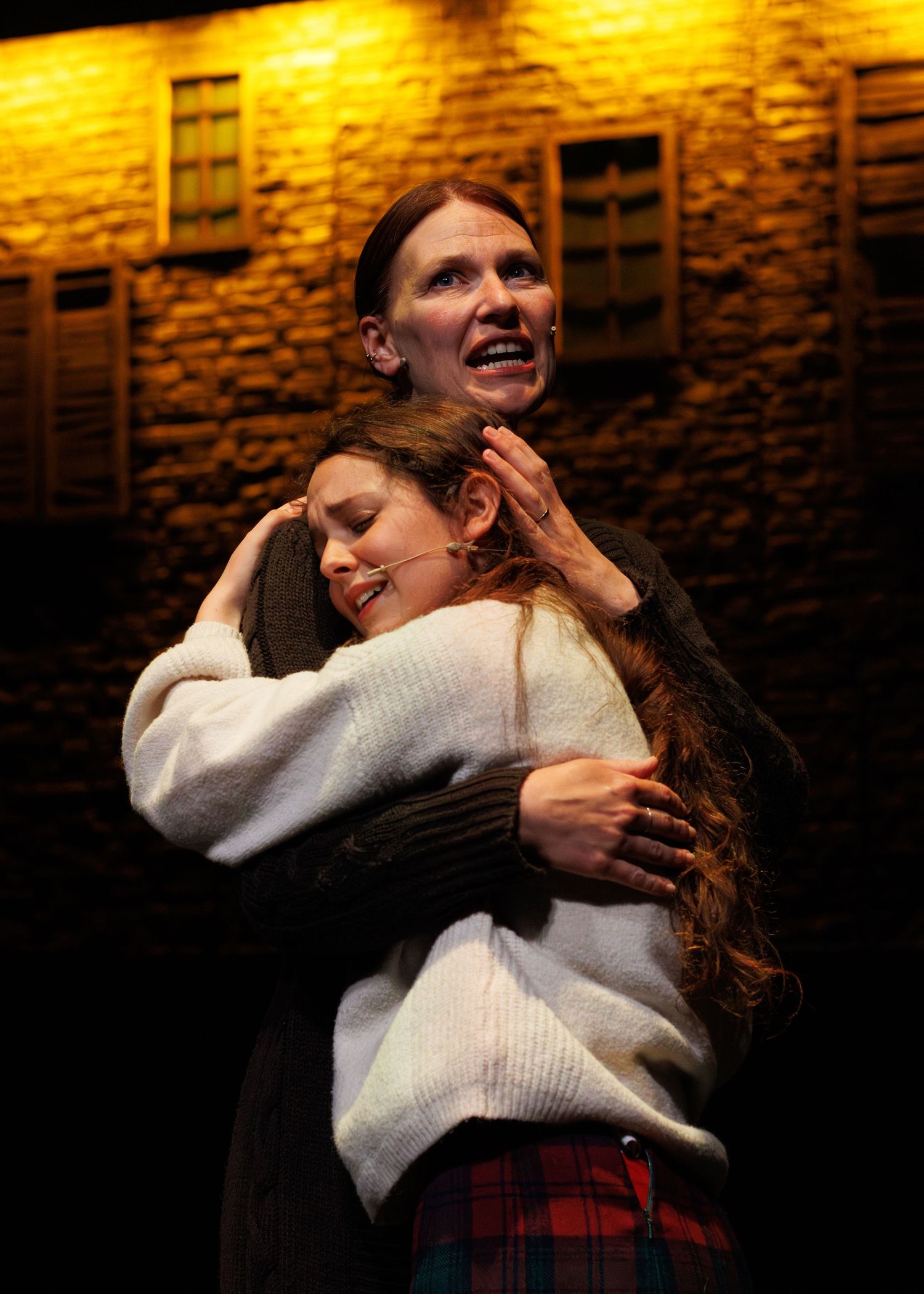 A bittersweet emotional moment shows two people sharing an embrace against a warm brick wall with dramatic lighting.