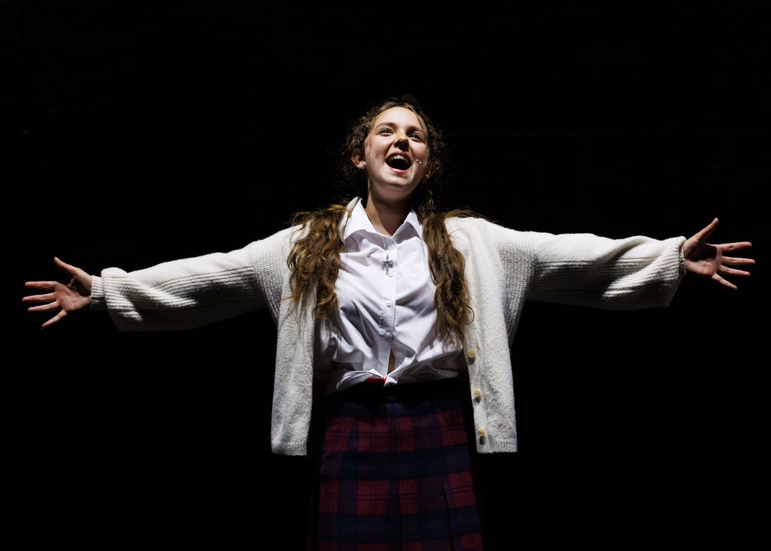 A performer in a white jacket and plaid skirt stands with arms outstretched against a dark background on stage.