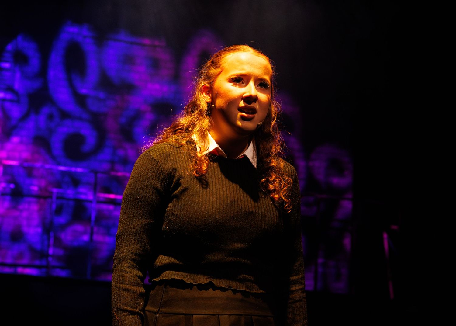Performer in black sweater sings emotively against purple-lit decorative backdrop on dark stage.