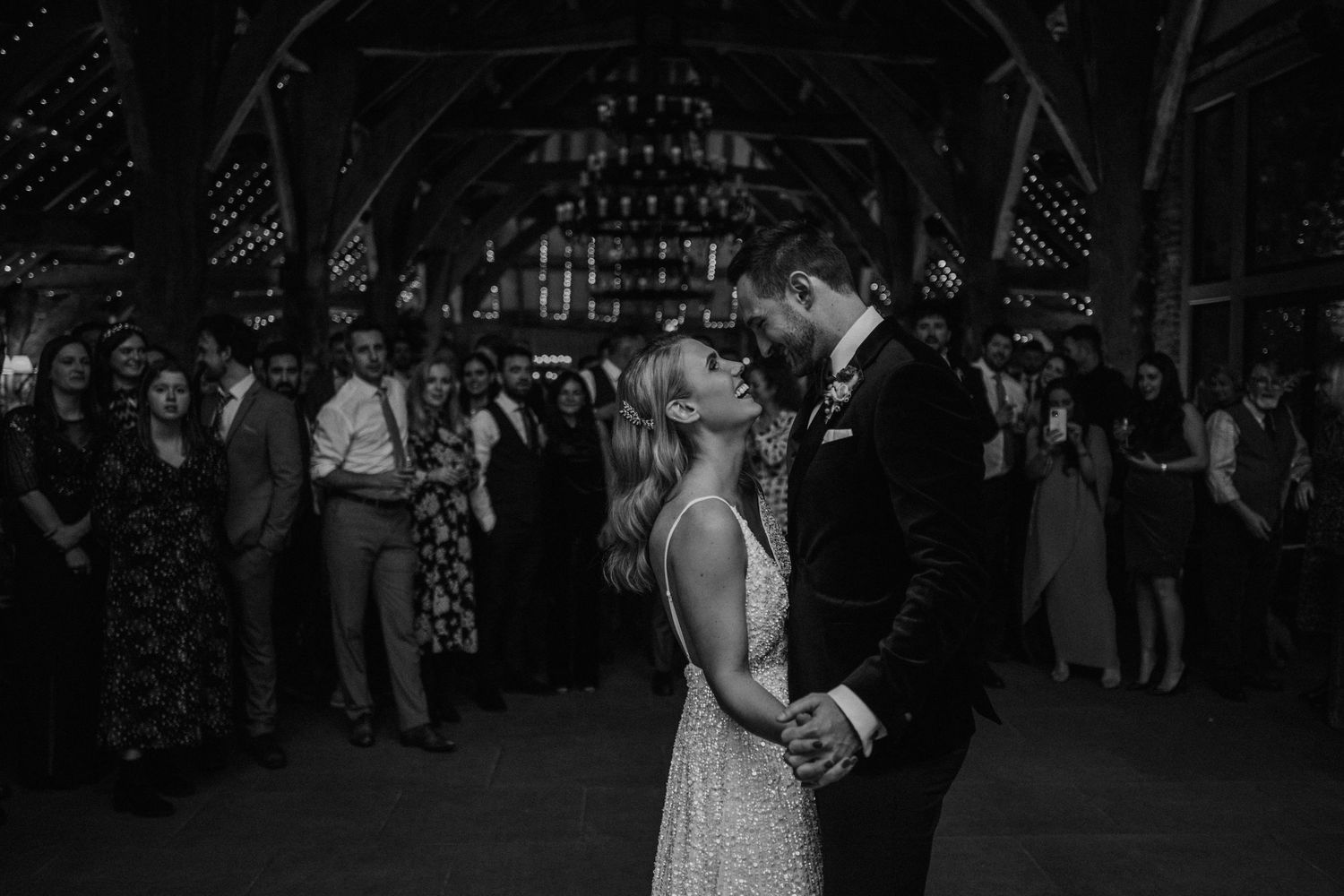 A couple shares a romantic dance at a dark rustic barn venue while guests watch from the sidelines.